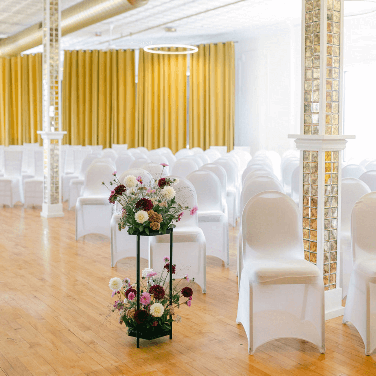 two tiered floral stand for a wedding surrounded by oak floors white covered chairs and golden curtains